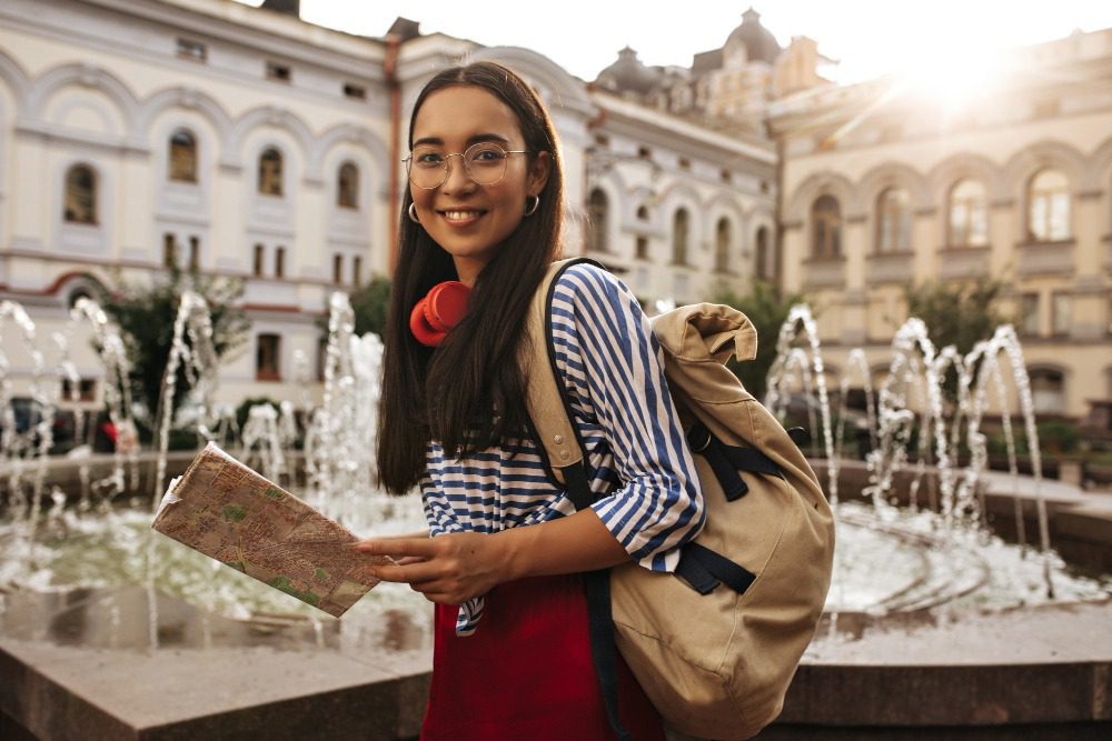  Jovem estudante com mochila, segurando um mapa e sorrindo, na frente de fonte de água em uma praça histórica, durante o dia.