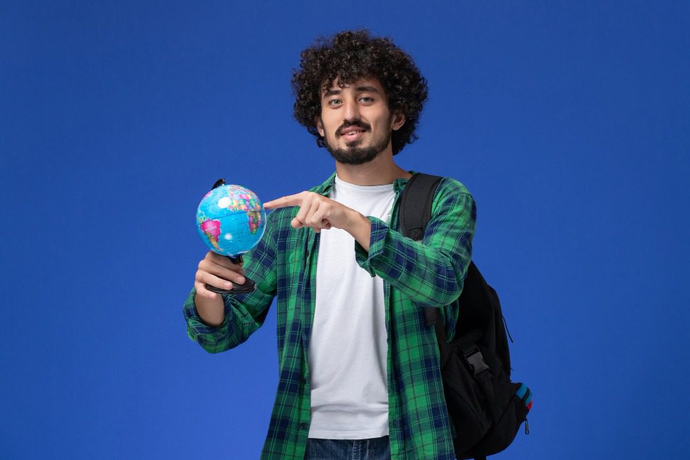 Jovem estudante com cabelo cacheado, mochila nas costas e camiseta branca, apontando para um globo terrestre em fundo azul, simbolizando a importância da educação e da curiosidade pelo mundo.
