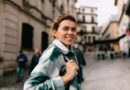 Mulher sorridente caminhando por uma rua de cidade antiga, usando jaqueta de xadrez e mochila, em um dia ensolarado com edifícios históricos ao fundo.