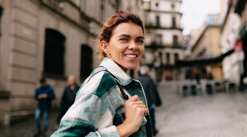 Mulher sorridente caminhando por uma rua de cidade antiga, usando jaqueta de xadrez e mochila, em um dia ensolarado com edifícios históricos ao fundo.
