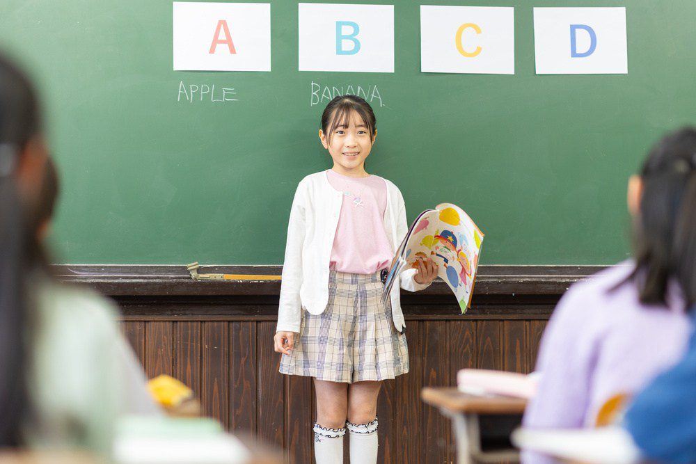 Professora ensinando alunas na sala de aula com quadro-negro ao fundo, destacando letras A, B, C e D e palavras como 'Apple' e 'Banana'