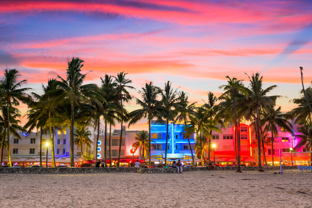 Imagem de um cenário costeiro ao pôr do sol com palmeiras e edifícios iluminados em cores vibrantes, refletindo uma atmosfera tropical e moderna.

