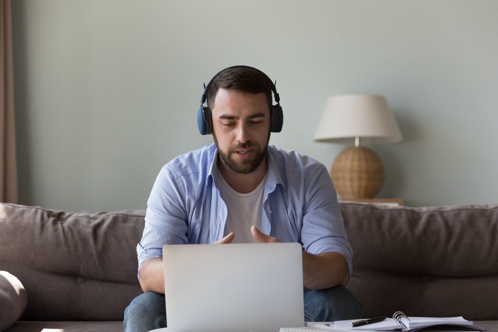 Homem com barba usando fones de ouvido e estudando inglês durante aula ao vivo, em ambiente confortável com iluminação natural.