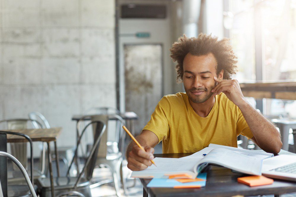 Homem estudando com livros e anotações em uma cafeteria moderna, aprendendo e se preparando para exames ou trabalhos acadêmicos.